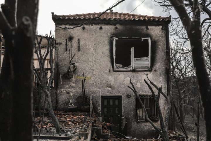 Ein durch Brand stark beschädigtes Wohnhaus mit verkohlter Fassade und zerstörten Fenstern zwischen verbrannten Bäumen.