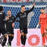 Bayer-Leverkusen-Spieler feiern vor dem Tor, während der gegnerische Torhüter und ein Verteidiger zuschauen.