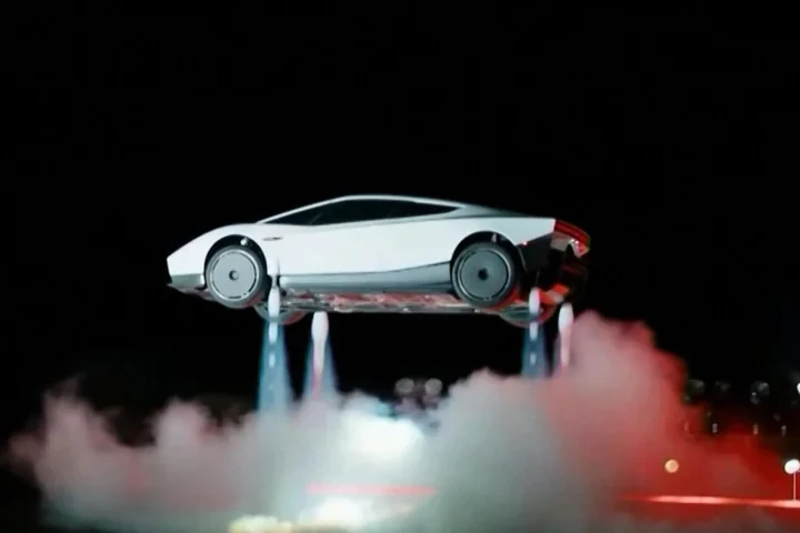 Ein weißer Tesla Roadster schwebt mit sichtbaren Raketendüsen über einer Bühne in einer dunklen Szene.