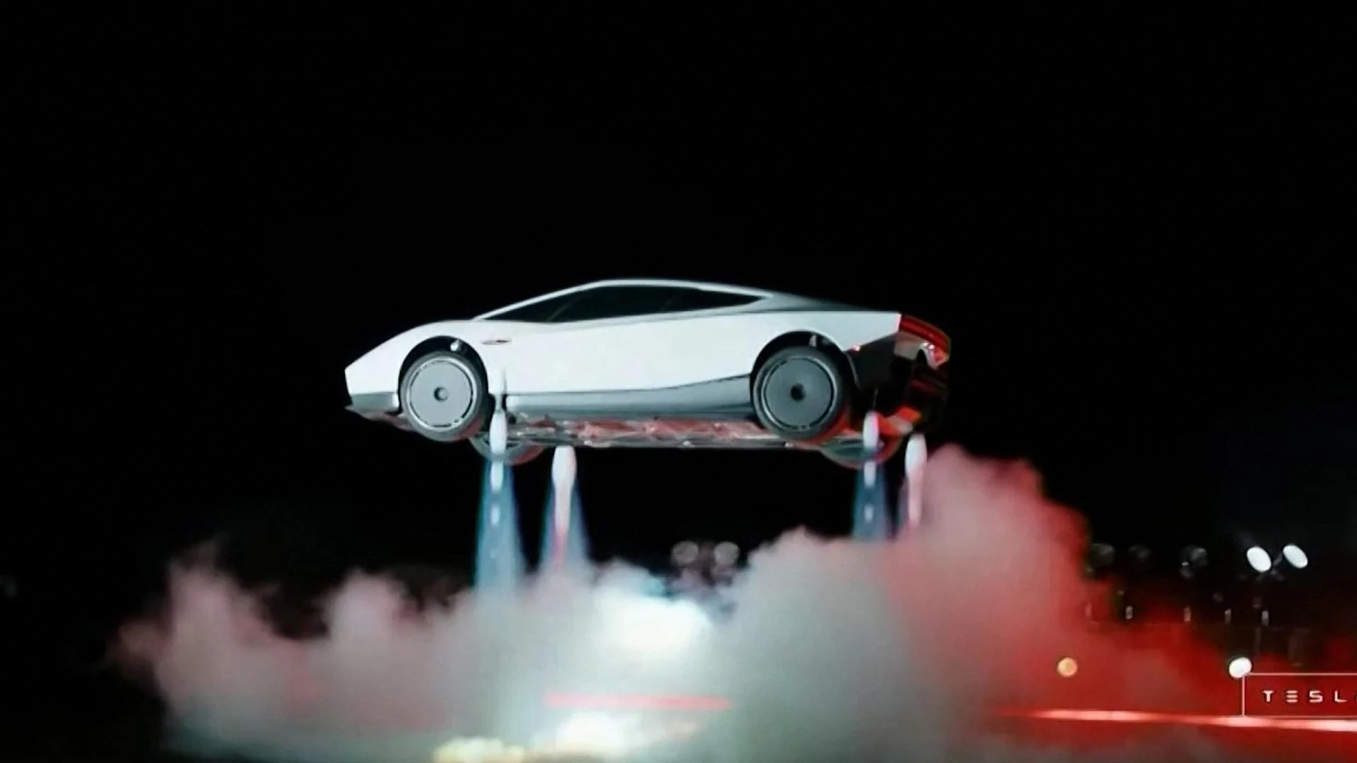 Tesla zeigt fliegenden Roadster in Waco und nennt Produktionsstart für 2028 1 Ein weißer Tesla Roadster schwebt mit sichtbaren Raketendüsen über einer Bühne in einer dunklen Szene.