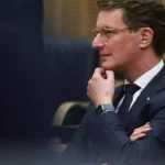 Hendrik Wüst sitzt in einem Sitzungssaal und blickt nach vorn, die Hand am Kinn.
