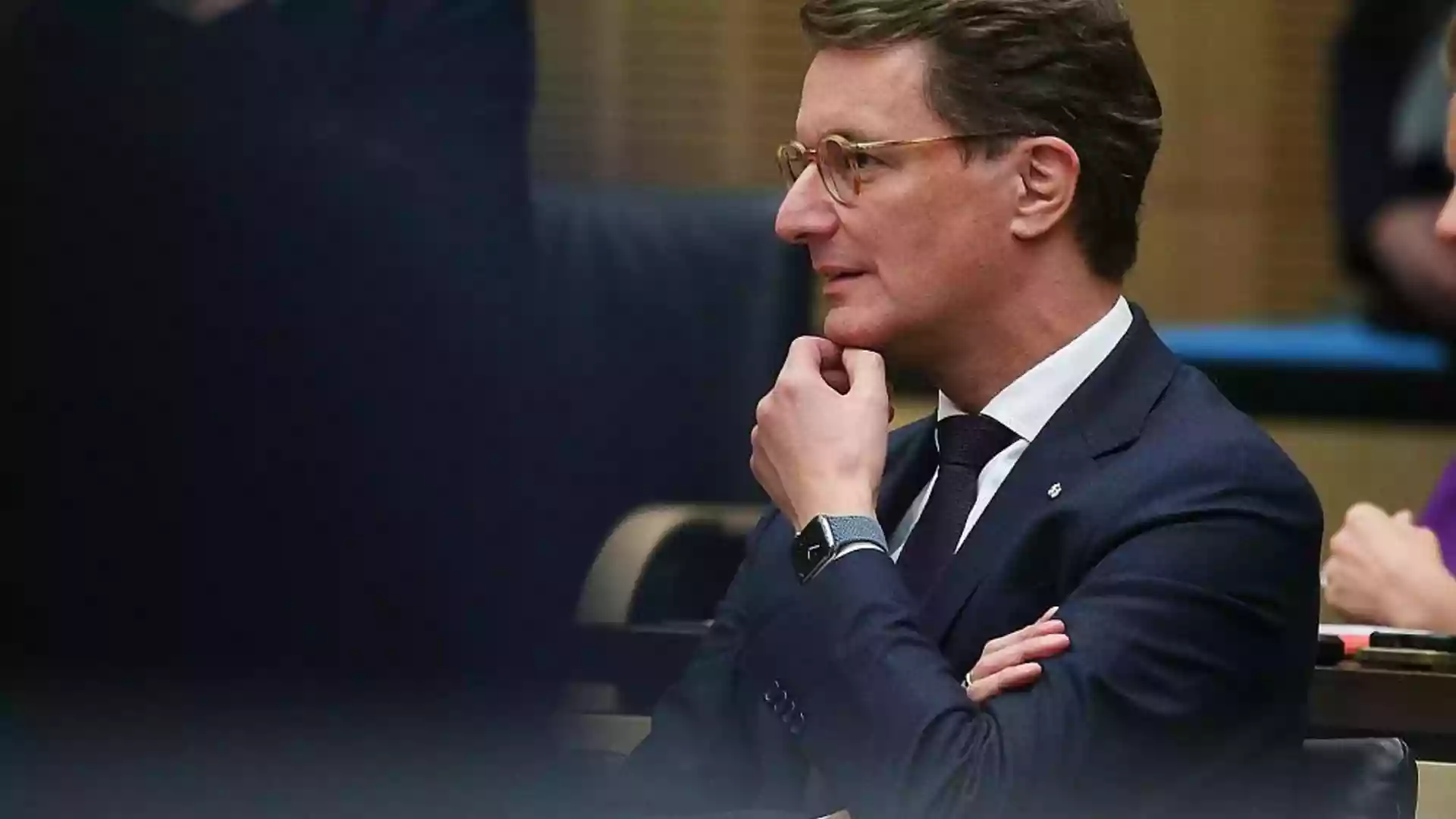 Hendrik Wüst sitzt in einem Sitzungssaal und blickt nach vorn, die Hand am Kinn.