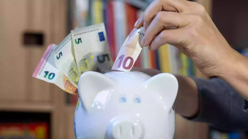 Eine Hand steckt einen 10-Euro-Schein in ein Sparschwein, daneben liegen weitere Euro-Banknoten.