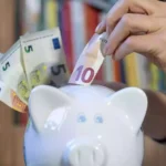 Eine Hand steckt einen 10-Euro-Schein in ein Sparschwein, daneben liegen weitere Euro-Banknoten.