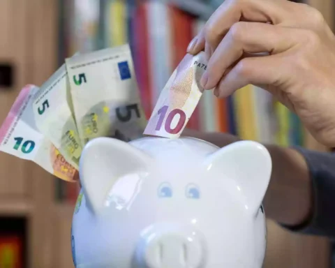 Eine Hand steckt einen 10-Euro-Schein in ein Sparschwein, daneben liegen weitere Euro-Banknoten.