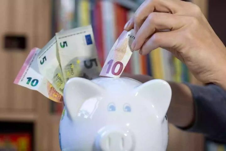 Eine Hand steckt einen 10-Euro-Schein in ein Sparschwein, daneben liegen weitere Euro-Banknoten.