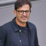 Ein Mann mit Brille und dunkler Jacke vor grauem Hintergrund.