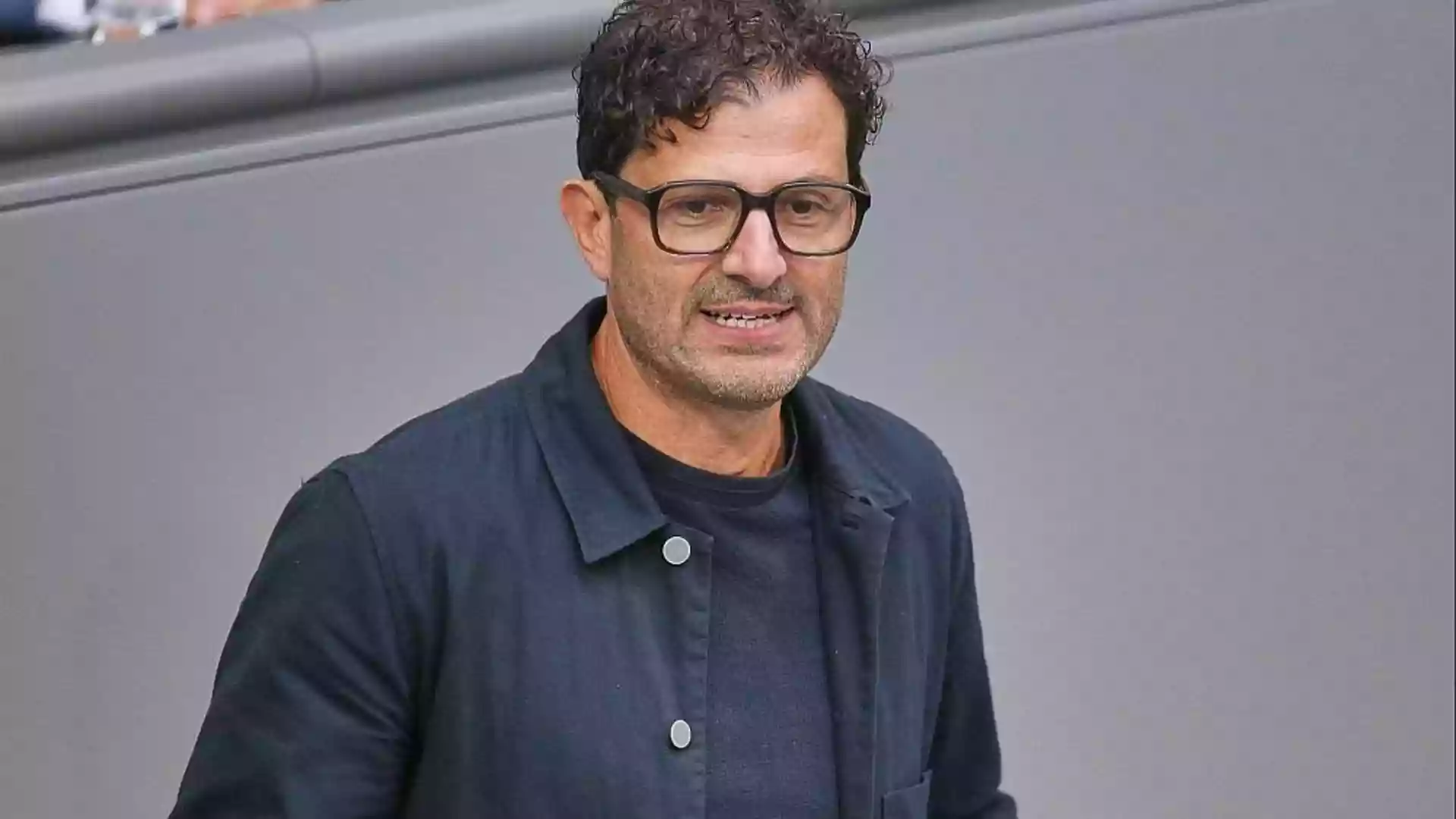 Ein Mann mit Brille und dunkler Jacke vor grauem Hintergrund.