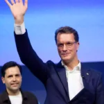 Hendrik Wüst winkt auf einer Bühne vor blauem Hintergrund.