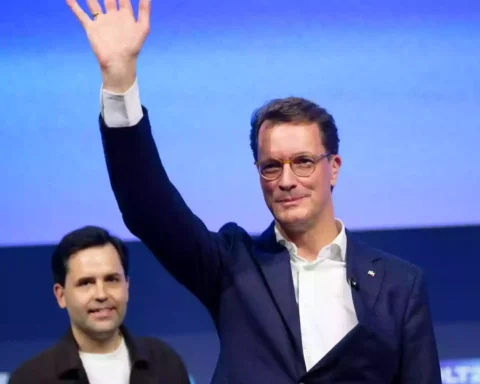Hendrik Wüst winkt auf einer Bühne vor blauem Hintergrund.