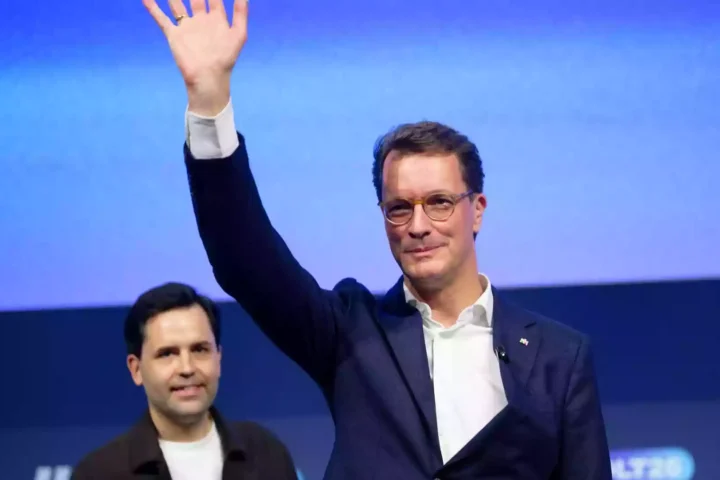 Hendrik Wüst winkt auf einer Bühne vor blauem Hintergrund.